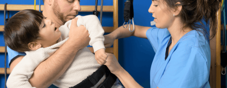 Obrazek wyróżniający w artykule rehabilitacja neurologiczna dzieci. Przedstawia ojca trzymającego niepełnosprawne dziecko i terapeutę