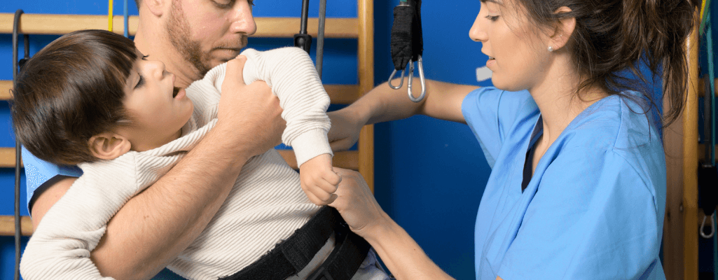 Rehabilitacja neurologiczna dzieci drogą do samodzielności. Skuteczne metody 1 Obrazek wyróżniający w artykule rehabilitacja neurologiczna dzieci. Przedstawia ojca trzymającego niepełnosprawne dziecko i terapeutę