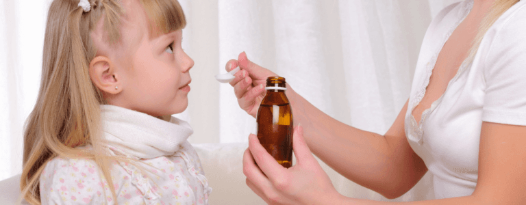 Obrazek wyróżniający w artykule syrop Junior angin. Przedstawia matkę podającą swojej córce syrop nalany z butelki na łyżeczkę