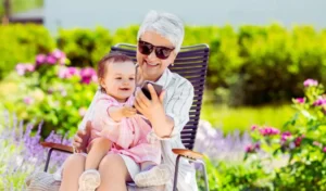 Obrazek wyróżniający do artykułu aktywny rodzic. Na zdjęciu widzimy starszą kobietę, prawdopodobnie babcię, siedzącą na leżaku w ogrodzie. Na kolanach trzyma małe dziecko, które z zainteresowaniem patrzy na smartfon, który kobieta trzyma w ręce. Oboje uśmiechają się, a dziecko wyciąga rękę w stronę telefonu, jakby chciało go dotknąć. Kobieta ma krótkie, siwe włosy i nosi ciemne okulary przeciwsłoneczne oraz jasną, prążkowaną koszulę. Scena rozgrywa się w pięknym, zielonym ogrodzie, otoczonym kwitnącymi kwiatami, co nadaje zdjęciu pogodny, ciepły charakter.