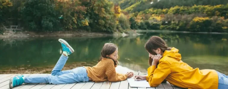 Obrazek wyróżniający do artykułu gdzie na majówkę z dziećmi. Widoczny 2 dziewczynki czytające książki na pomoście nad jeziorem. W tle widoczny zarys zielonych gór.