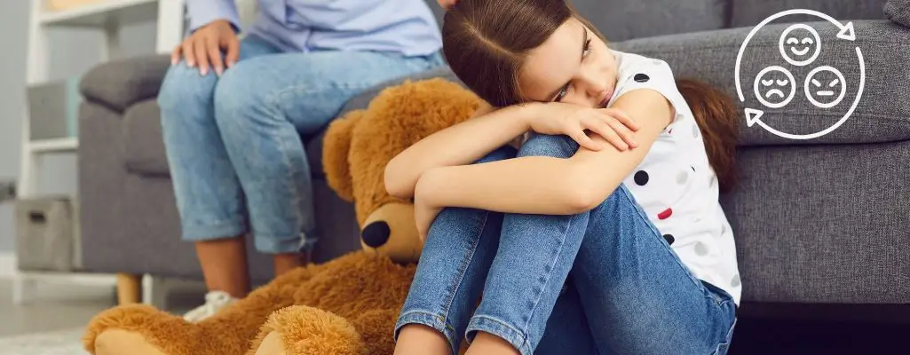 Emocje dziecka. Jak je rozpoznawać i właściwie reagować? 1 Obrazek do artykułu - emocje dziecka. Obrazek przedstawia młodą dziewczynę siedzącą na podłodze, opartą o kanapę, obejmującą kolana i wyglądającą na smutną. Obok niej znajduje się duża pluszowa maskotka w kształcie misia. W tle widoczne są dwie osoby (dorosły i dziecko) siedzące na kanapie, skupione na czymś innym, poza kadr obrazu. W prawym górnym rogu obrazka znajduje się symbol z trzema różnymi emocjami twarzy: szczęście, neutralność i smutek. Ten obraz może oddawać uczucia osamotnienia lub smutku dziecka, pomimo obecności innych osób w pokoju, sugerując subtelne trudności w komunikacji emocjonalnej w rodzinie.