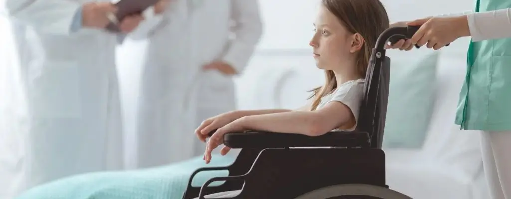 Orzeczenie o niepełnosprawności dziecka. Przewodnik dla rodziców i opiekunów 1 Obrazek wyróżniający do artykułu - orzeczenie o niepełnosprawności dziecka. Zdjęcie przedstawia młodą dziewczynkę siedzącą na wózku inwalidzkim. Dziewczynka ma brązowe włosy, które są proste i sięgają ramion. Ma na sobie białą bluzkę z krótkim rękawem. W tle widać dwóch lekarzy, którzy wydają się być zaangażowani w rozmowę lub przeglądanie dokumentów. Jeden z lekarzy stoi, a drugi jest częściowo widoczny. Dziewczynkę pcha osoba, której ręce są widoczne, ubrana w biały uniform medyczny. Tło jest jasne, co może sugerować, że zdjęcie zostało wykonane w placówce medycznej, takiej jak szpital lub klinika. Dziewczynka patrzy przed siebie z poważnym wyrazem twarzy.