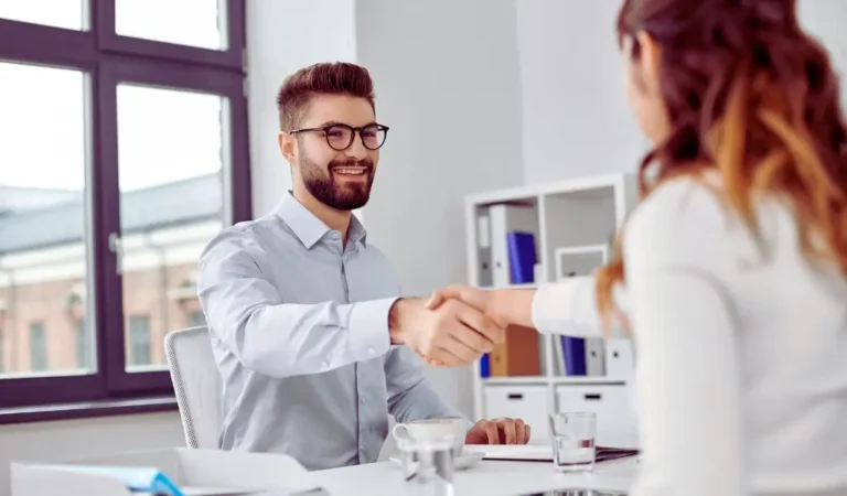 Obrazek wyróżniający do artykułu - słabe strony podczas rozmowy kwalifikacyjnej. Na zdjęciu widoczna jest scena w biurze, gdzie mężczyzna w okularach i z brodą podaje rękę kobiecie, siedzącej naprzeciwko niego. Mężczyzna ma na sobie jasnoniebieską koszulę i wygląda na zadowolonego, uśmiecha się serdecznie. Kobieta jest częściowo widoczna, siedzi tyłem do kamery, a jej ręka jest wyciągnięta do uścisku. Scena sugeruje zakończenie rozmowy kwalifikacyjnej lub innego spotkania biznesowego, na którym doszło do porozumienia lub pozytywnego wyniku. W tle widać okno oraz regał z dokumentami, co podkreśla biurowy charakter tego miejsca.