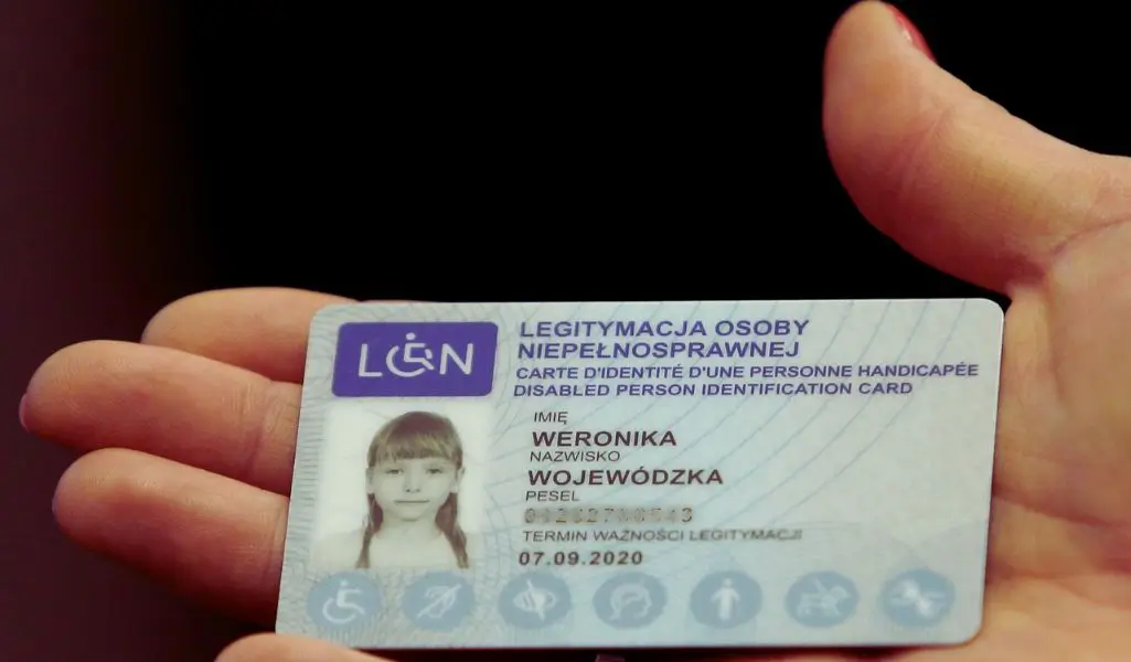 Co warto wiedzieć o legitymacji osoby niepełnosprawnej? 1 Na zdjęciu widoczna jest Legitymacja osoby niepełnosprawnej, trzymana w dłoni. Dokument ma jasnoniebieskie tło z subtelnymi wzorami i symbolami wskazującymi na różne rodzaje niepełnosprawności. W lewym górnym rogu znajduje się niebieski znak z symbolem wózka inwalidzkiego i literami "LON". W centralnej części znajduje się czarno-białe zdjęcie młodej dziewczynki o jasnych włosach, z grzywką, patrzącej spokojnym wzrokiem. Dane osobowe, takie jak imię, nazwisko oraz PESEL (częściowo ukryty), są wydrukowane w wyrazistej czcionce. Poniżej znajduje się termin ważności dokumentu, który upłynął 7 września 2020 roku.