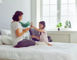 Na zdjęciu widać mamę i córkę siedzące naprzeciwko siebie na łóżku w jasnym, przytulnym pokoju. Bawią się wspólnie w grę „klaszczącą” – obie uśmiechają się szeroko, patrząc sobie w oczy, co podkreśla ich bliskość i radość z bycia razem. W tle widać duże okno z doniczkami na parapecie, przez które wpada naturalne światło. Obrazek oddaje ciepło domowej atmosfery i pokazuje, czym są dobre relacje z dzieckiem – pełne uważności, zabawy i wzajemnego zrozumienia.