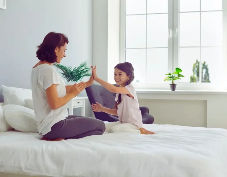 Na zdjęciu widać mamę i córkę siedzące naprzeciwko siebie na łóżku w jasnym, przytulnym pokoju. Bawią się wspólnie w grę „klaszczącą” – obie uśmiechają się szeroko, patrząc sobie w oczy, co podkreśla ich bliskość i radość z bycia razem. W tle widać duże okno z doniczkami na parapecie, przez które wpada naturalne światło. Obrazek oddaje ciepło domowej atmosfery i pokazuje, czym są dobre relacje z dzieckiem – pełne uważności, zabawy i wzajemnego zrozumienia.