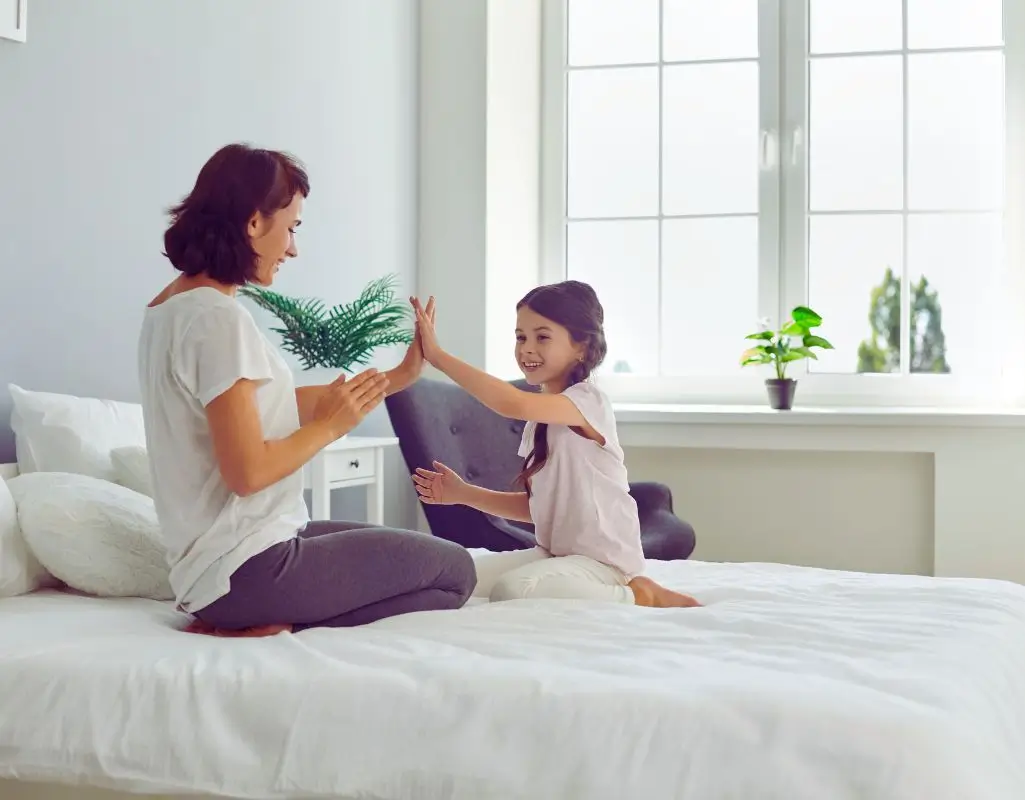 Odkryj 10 fundamentów, na których zbudujesz dobre relacje z dzieckiem i je zachowasz 4 Na zdjęciu widać mamę i córkę siedzące naprzeciwko siebie na łóżku w jasnym, przytulnym pokoju. Bawią się wspólnie w grę „klaszczącą” – obie uśmiechają się szeroko, patrząc sobie w oczy, co podkreśla ich bliskość i radość z bycia razem. W tle widać duże okno z doniczkami na parapecie, przez które wpada naturalne światło. Obrazek oddaje ciepło domowej atmosfery i pokazuje, czym są dobre relacje z dzieckiem – pełne uważności, zabawy i wzajemnego zrozumienia.