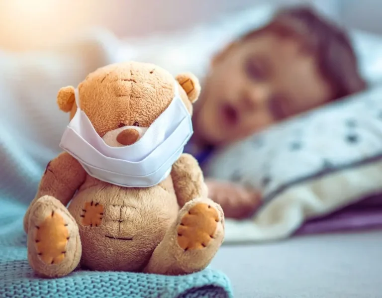 Na zdjęciu widać pluszowego misia siedzącego na kocu, z założoną maseczką ochronną na pyszczku. W tle, lekko rozmytym, leży na poduszce śpiące dziecko — wygląda na chore lub zmęczone, przytulone do pościeli. Obrazek budzi emocje troski, czułości i współczucia. Przedstawia codzienność wielu rodzin, gdzie choroba dziecka wymaga opieki i wsparcia. Pluszowy miś z maseczką symbolizuje zarówno dbałość o zdrowie, jak i dziecięce poczucie bezpieczeństwa. Można go użyć jako ilustrację do treści związanych z tematem: lista chorób do bezterminowego orzeczenia.