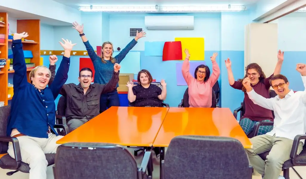 Czy Warsztaty Terapii Zajęciowej to dobre miejsce dla Twojego dziecka? Praktyczny przewodnik dla opiekuna 3 Na zdjęciu widać grupę osób siedzących przy stole w jasnym, kolorowym pomieszczeniu. Wszyscy mają podniesione ręce i uśmiechy na twarzach — emanują radością, entuzjazmem i poczuciem wspólnoty. W tle znajdują się kolorowe kartki na ścianach oraz półki z materiałami, co wskazuje na kreatywną przestrzeń. To moment pełen pozytywnej energii, prawdopodobnie uchwycony podczas wspólnej aktywności lub świętowania w miejscu takim jak Warsztat Terapii Zajęciowej. Obrazek oddaje atmosferę wsparcia, akceptacji i zaangażowania — pokazuje, jak ważna jest integracja i wspólne przeżywanie sukcesów.