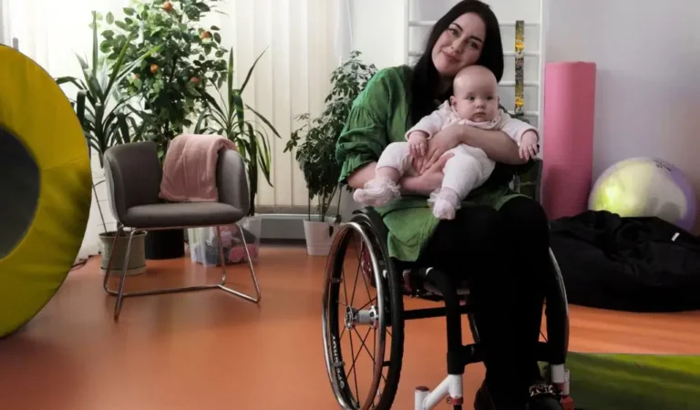 Kobieta na wózku inwalidzkim trzymająca niemowlę na kolanach w domowym otoczeniu – dofinansowanie do opieki nad dzieckiem.